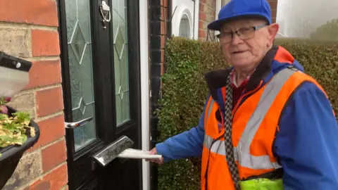 A man in his 70s looks at the camera while putting a newspaper through the letter box of a black front door. He is wearing glasses, a blue cap and blue jacket with a flourescent orange high-vis vest.