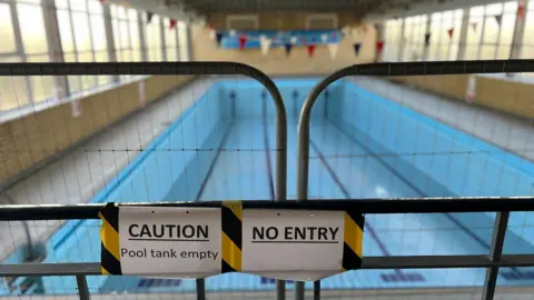 Ann Gannon / BBC A dry, empty swimming pool is cordoned off by metal fencing. Two signs on the barrier read “Caution: Pool tank empty” and “No entry”. The blue-tiled pool stretches the length of the hall, with lane markings visible on the floor. Large windows line the left side, and bunting hangs above the far end of the pool.
