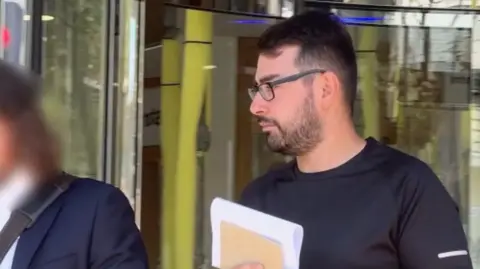 BBC A man with short dark hair and glasses walks past a glass-fronted building entrance while holding a folder and papers. Another individual appears partially blurred in the foreground on their left side. The background shows reflective glass panels and light-colored interior fixtures visible through the doorway.