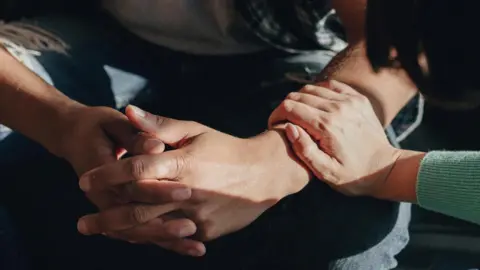 Generic close up image of a man sitting down with his fingers interlocked while a woman rests her hand on his forearm in a supportive way