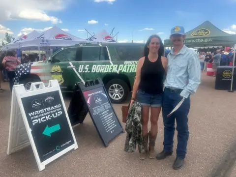 A young man in a baseball hat stands in front of a tent advertising the Customs and Border Patrol, with his mom standing next to him in cowboy boots