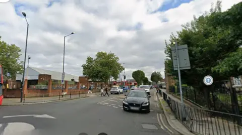 Google street view image of Pound Lane from its junction with High Road, showing a road with several cars, pedestrians and what looks like a large grocery store to the left.