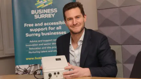 Surrey County Council The image shows a man smiling for a photograph, while holding a white electrical object. He is wearing a navy blue suit with a striped undershirt. There is a blue banner behind him that reads: "Business Surrey, Free and accessible support for all Surrey businesses" with some smaller text underneath.