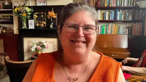 Sam Read/BBC Woman with orange sleeveless top and a gold necklace. She is smiling and is sting in a pub with bookshelves,a piano and a fireplace behind her.