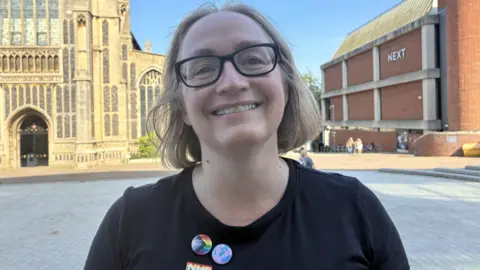 Zoe Dennis/BBC A woman smiles at the camera in a head and shoulders image. She is wearing black framed spectacles and a black tshirt with a Progress Pride pin badge on it, made up of rainbow stripes and an arrow made up of black, blue, pink and white stripes. She has blonde hair in a bob. 