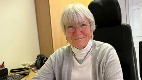 A woman with grey hair in a white top and pale grey cardigan, sitting on a high-backed black chair at a wooden desk with a black keyboard on it