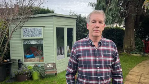 Andrew Causley in a blue, red and white check shirt standing in front of his light green summerhouse which has planters in front of it in his garden.