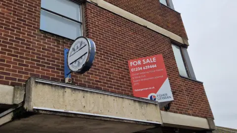 A disused police station. The red brick building has a Northamptonshire Police sign on it, which has been taped over and a "for sale" sign.