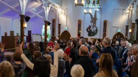 St Barbara's A congregation gathers in a church with white walls and wooden beams supporting arches which reach towards the ceiling. It is lit by lamps on the walls and hanging from above, and members of the congregation have their hands in the air.