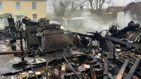 Andrew Turner/BBC The wreckage of a fire outside a house. Burnt remains can be seen at the front of the building. Smoke can also be seen coming off of the wreckage. 