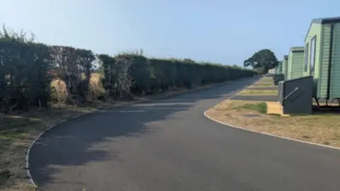 A row of green static caravans can be seen on the right hand side of the image. There is a sweeping tarmac road going past them in the centre of the image with a hedge on the left.