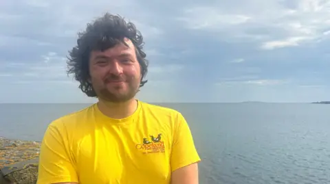 Adam has thick curly hair that is blowing in the wind. He has dark stubble and wears a yellow t-shirt. Behind him is the sea, sky and horizon.