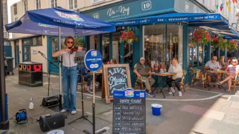 A violinist performs outside a restaurant on a sunny day during the Guernsey Street Festival. She is standing in front of an iPad while she performs and is wearing a white T-shirt, blue jeans, white trainers and sunglasses. People are sitting at various tables outside the restaurant and they are watching her.