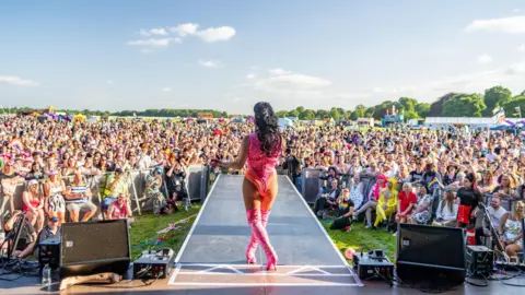 York Pride / Milner Creative A drag performer in a pink and orange leotard with pink stiletto boots performs in from of a crowd of hundreds of people.