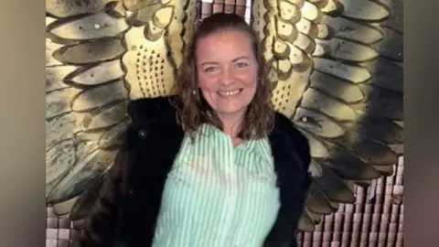 Dianne Cleary pictured in front of a wall with a large bronze set of angel wings on it. She is smiling and wearing a stripey green and white shirt and open black coat. Her wavy hair is half pinned back from her face.