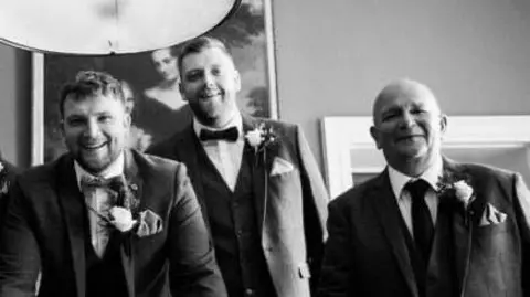 Sam, Olly and Phil all smile into the camera. They're wearing matching suits for a wedding, with flowers in their lapels. The photo is black and white. 