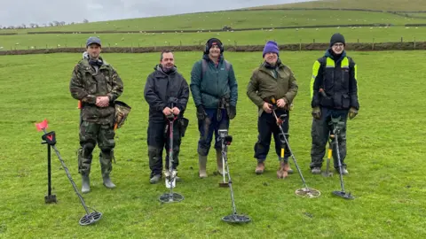 Five men are standing in an open field, all dressed in outdoor clothes, wearing jackets, boots and hats. Each is holding a metal detector, with two other detectors resting on the ground in front of them. The backdrop is a grassy, rolling landscape, with fences and a few scattered livestock distant in the background.