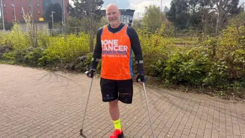 Peter Lloyd, who had his left leg amputated, is posing for a picture with his crutches. He is wearing a Bone Cancer Research Trust vest. 