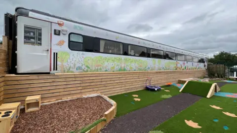 Stuart Woodward/BBC A converted railway carriage which has been turned into classroom space for a pre-school. Outside the carriage there is a sensory garden with astro turf, an area covered in wood chippings, and colourful symbols on the ground.