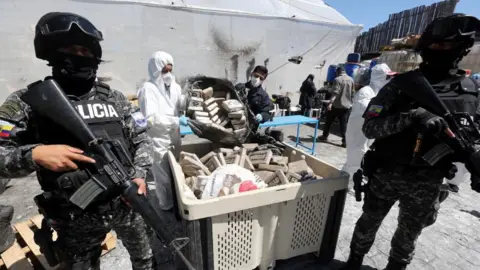 Polisi Reuters dengan senjata berjaga di atas sebuah wadah yang diisi dengan paket narkoba karena dua orang di topeng dan pakaian bahaya memberi lebih banyak obat di dalamnya