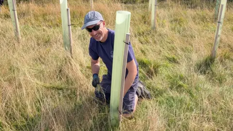 NECF Forest Manager Lloyd Jones crouches behind a sapling that has just been planted. He is wearing a dark t-shirt and shorts, baseball cap and sunglasses and is smiling. The sapling has a semi-translucent, green plastic tree guard around it and in the background there are several other newly-planted trees.