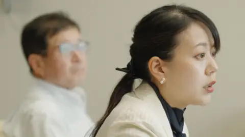 Shoko Kawata, a female mayor in a white blazer and dark blouse sits on an armchair. Her deputy in a light-coloured shirt is visible behind her.