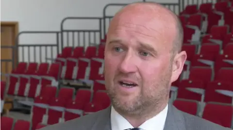 Police and Crime Commissioner Dafydd Llywelyn wearing a grey suit and stood in front of tiered seating