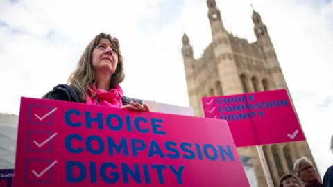 PA Media Humanists UK's members and supporters, during a protest outside the Houses of Parliament in London to call for reform as peers debate the new assisted dying legislation. A woman holds a bright pink placard reading "choice, compassion, dignity".