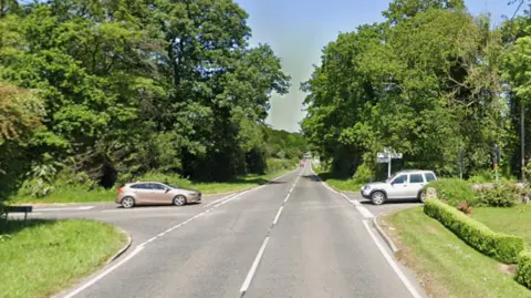 A junction is pictured with trees lining the road. Two cars can be seen waiting to cross on either side of the road.