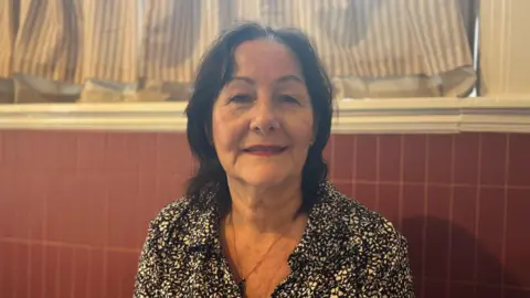 Janet with shoulder length black hair wearing a black and white patterned blouse sitting in front of a red-tiled wall and a window with light brown and white striped curtains. She is smiling.