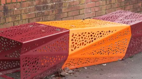 A metal bench which has no back to it in a misshapen cube shape with patterns in the metal. This one is in red and orange with a wall behind.