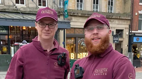 Two men wearing burgundy caps and fleeces stood next to each other. Both have black cameras attached to their chest. Jon, on the left, is clean shaven with glasses, while Scott has glasses and a red beard.