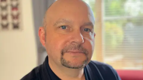 Lee Jackson, with blue eyes, a shaved head and a short beard and moustache, wearing a navy shirt.
