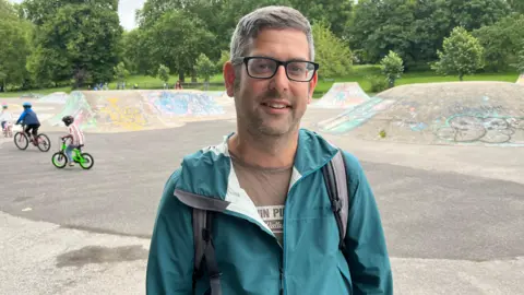Councillor Rob Bryher. He has short grey hair and is wearing a brown t-shirt under a green jacket. Children riding bikes near the skatepark can be seen behind him.