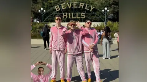 Boy Throb Three members of Boy Throb are standing in front of a Beverly Hills sign. They are all dressed in a pink velour tracksuit with white detailing. Darshan is photoshopped in to the bottom of the photo and he is holding his hands in a heart. 