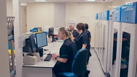 Four females nurses in uniform sit at desks at the hospital. They are sat in a vertical line in the photo with computers in front of them.
