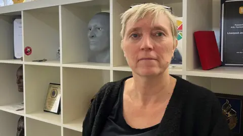 Professor Caroline Wilkinson has short blonde hair and is wearing a black t-shirt and black cardigan and is standing in front of a shelving unity which has sculptures of faces and a series of awards.