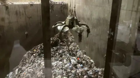 BBC News A pile of rubbish surrounded by large concrete walls. A large metal claw is hovering above the pile ready to pick it up.
