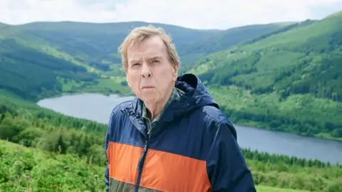 Timothy Spall in a waterproof jacket standing in the Bannau Brycheiniog/Brecon Beacons. There is a lake or reservoir behind him and hills around.