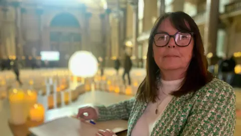 Barbara White stands beside the book with candles and paper lanterns glowing in the backdrop inside St George's Hall. She has shoulder-length brown hair and glasses and is wearing a green and white blazer.