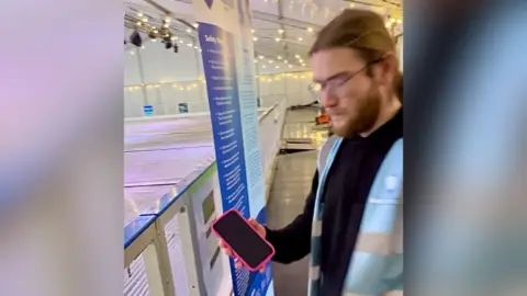 Destination MK An ice rink worker with long brown hair and glasses holds the pink iPhone next to the rink. He is wearing a black jumper and blue hi-vis jacket.