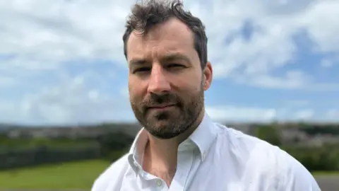 Gareth is wearing a white shirt and has brown hair and a beard. He is stood outside with the skyline and park area blurred in the background.