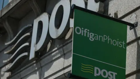 A green sign which says, in white writing, 'Oifig an Phoist'. Below that, there is a white line on the sign, and below that is written 'post', with four horizontal yellow lines to the left of it. The sign is in front of a brick building, which has a large stone sign saying 'post' on the front. 