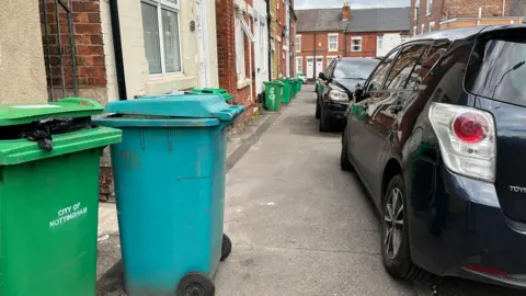 A green bin which says 'city of nottingham' on it, a blue bin and a black car partially on the pavement