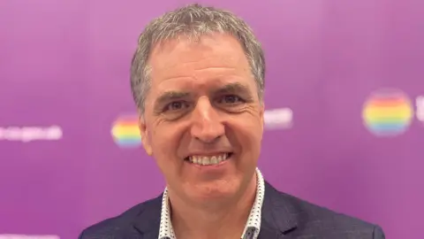 Claire Hamilton/BBC Steve Rotheram has greying hair, a charcoal grey jacket and white shirt with checks. It is a head shot of him and the background behind him is purple.