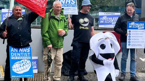 Six member of the Toothless in Suffolk campaign group stand outside Endeavour House. They are equipped with posters and flags.