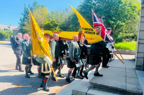 Coffin at funeral of veteran Jim Glennie being carried into a chapel by men in kilts, under a blue sky.