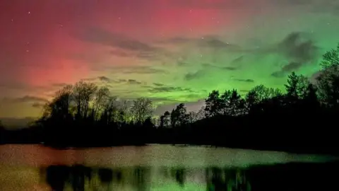 A pink and green glow in the night sky, reflected in a lake.