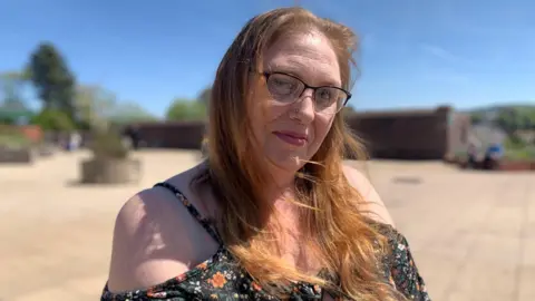 Melanie Harris in black summer dress with shoulder straps and floral print. She wears black-rimmed glasses and has long red hear worn loose. She is pictured outside in sunny weather