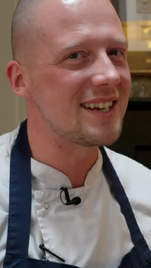 Luke Emmess grins at the camera. He is wearing chef whites and a blue apron over the top. He has light stubble on his chin and a shaved head.
